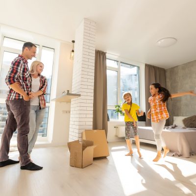 Excited young family have fun in living room unpacking cardboard boxes on moving day, happy parents play with little daughters jump out of carton package, overjoyed relocating to new home together
